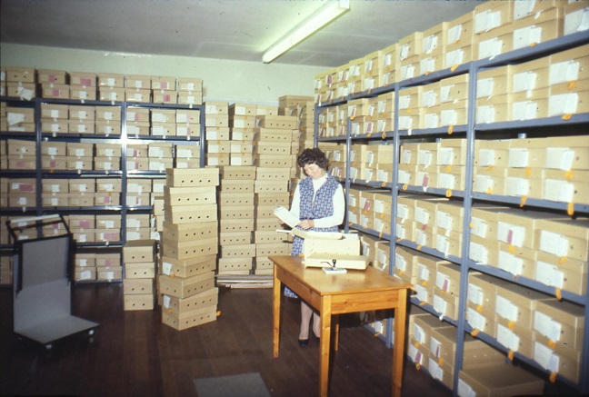 Modern Records Clerk standing at a small table, surrounded by document storage boxes on the floor and also on shelves