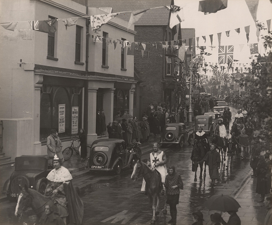 Black and white photograph of Horsham main street. Bunting and flags cross between the buildings. A line of men in mediaeval costumes ride on horses, with a helper holding the reins of the horses to keep them steady. It appears to be a wet day, with spectators holding umbrellas.